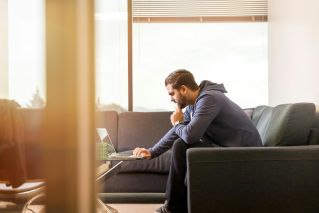 A man in casual attire works remotely from a sofa, using a laptop near a large window in a modern room.