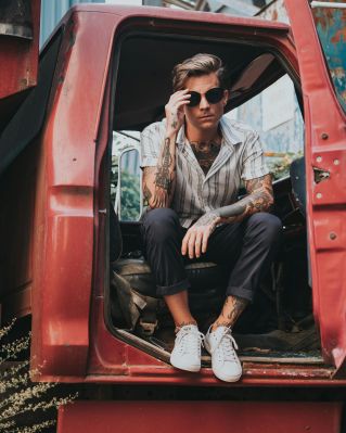 A tattooed man in sunglasses sits in a vintage truck, exuding modern style and cool attitude.